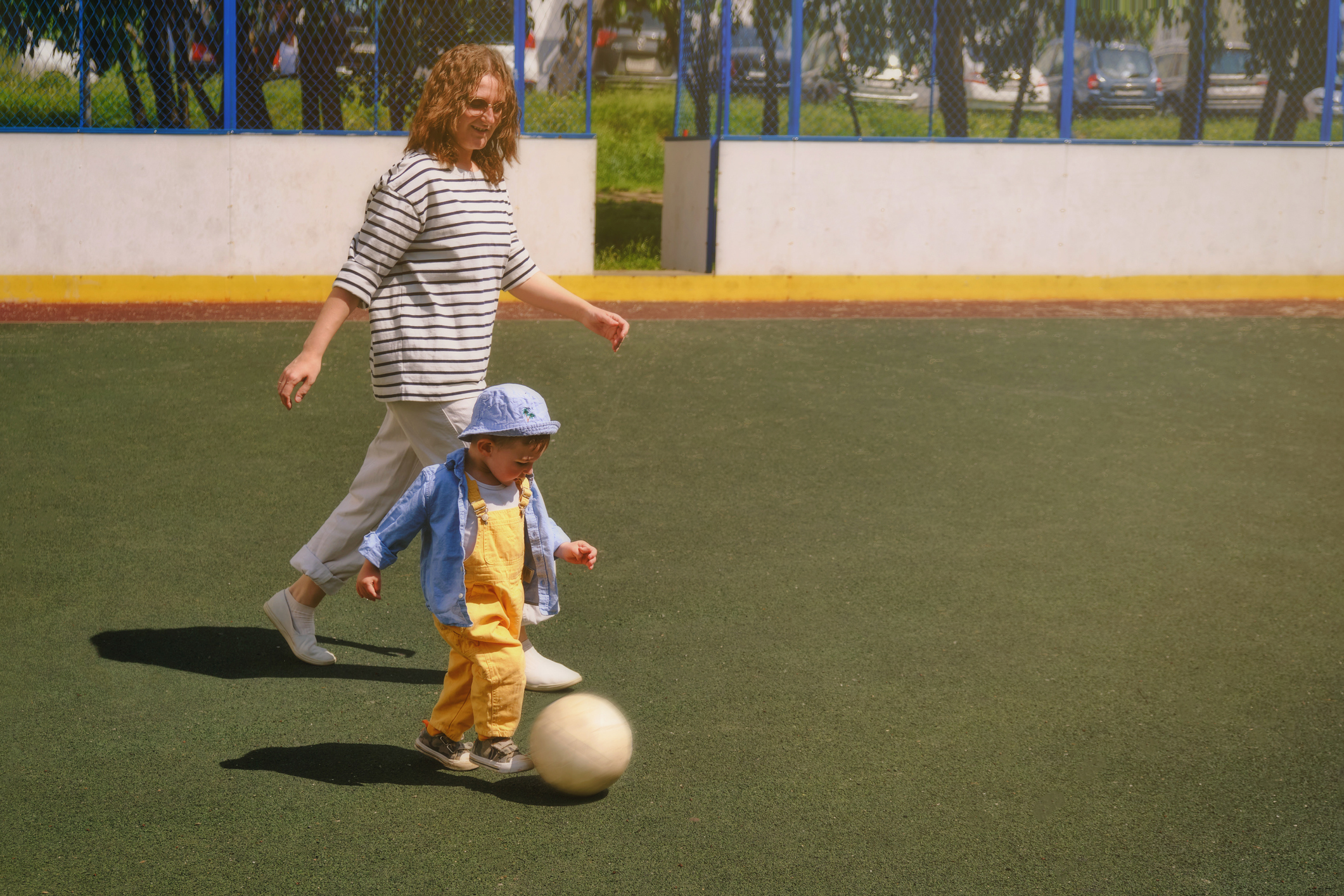 The child and woman mother is having fun kicking the soccer ball around on the outdoor field. Baby with mom is happily playing with the white ball in the park. Kid aged about two years.